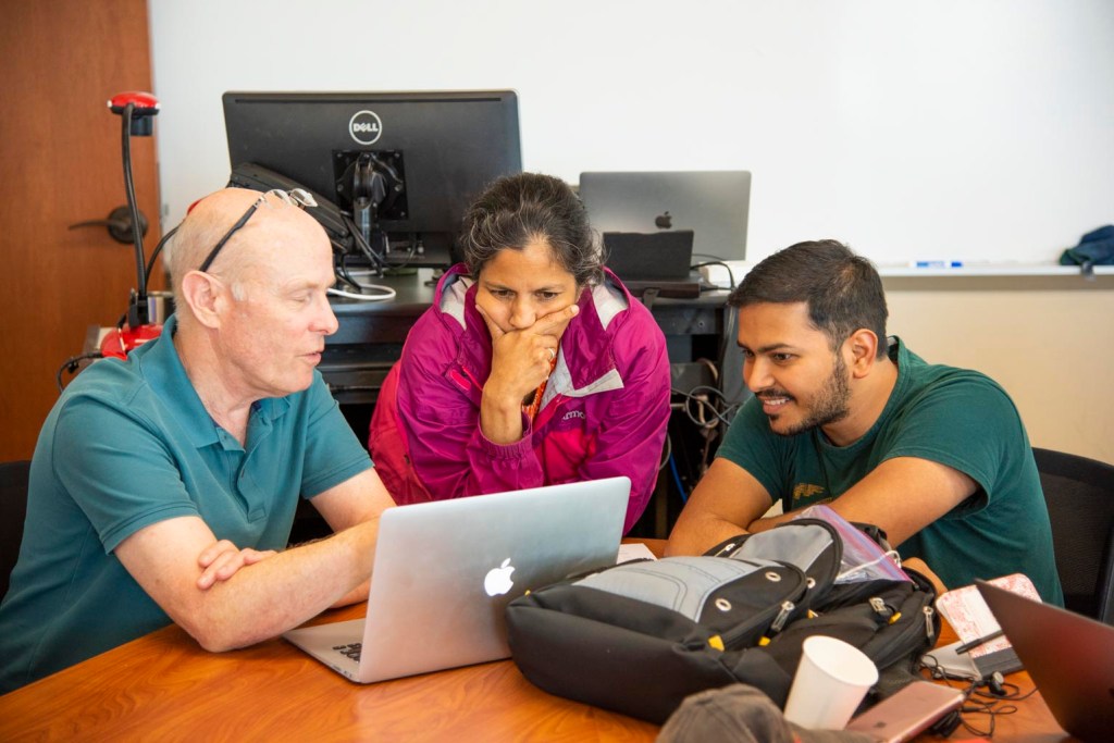 3 people stand around an Apple laptop looking at something on the screen. 