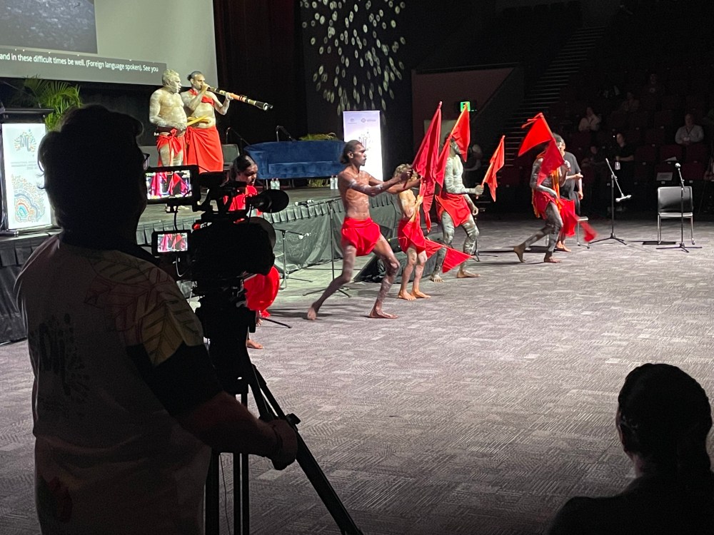 A group of men wearing red clothes and hold red flags, stand in front of a stange dancing