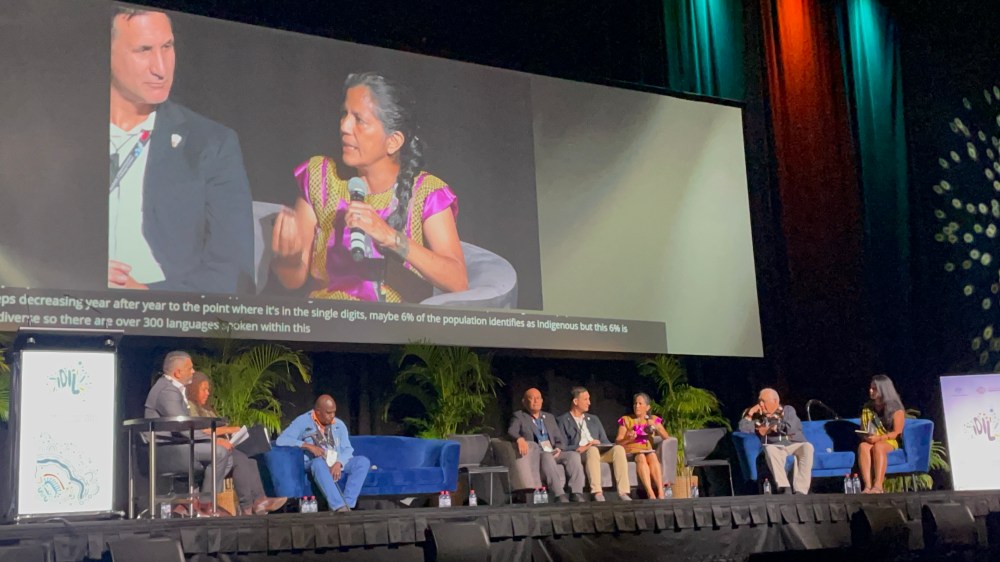 A group of people sit on a stage while a woman speaks into a microphone