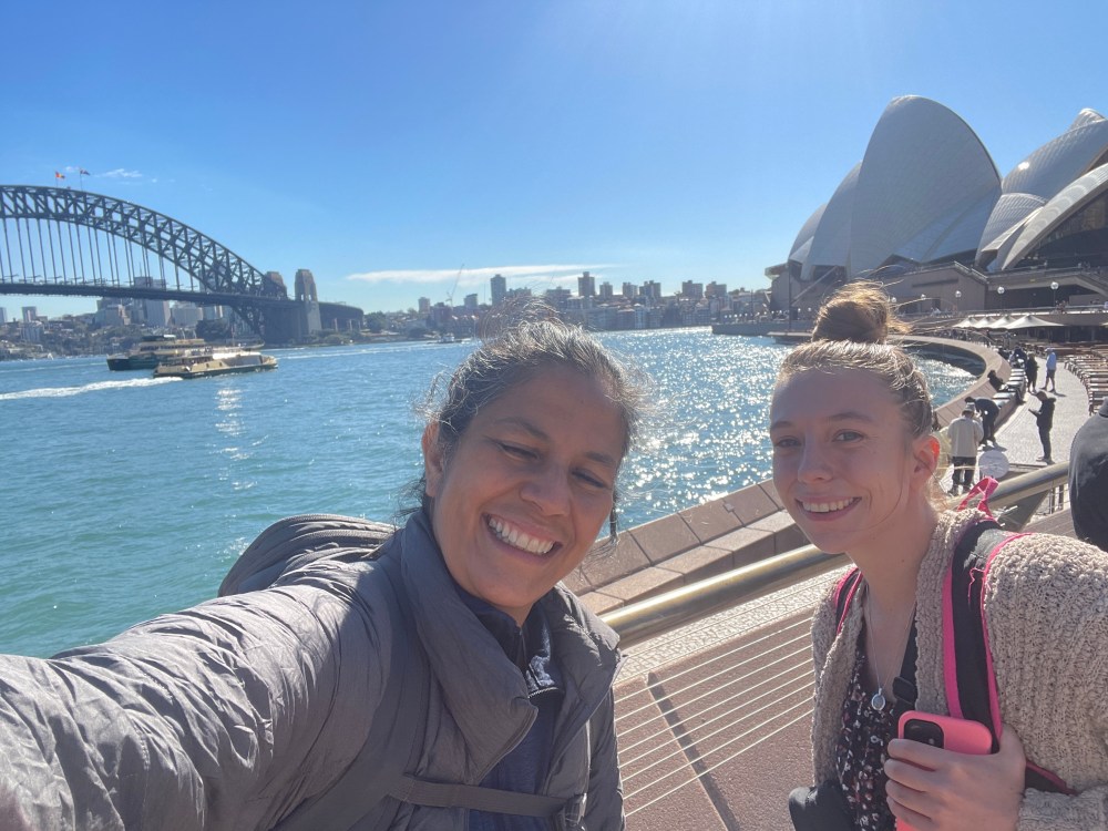 Two women stand in front of a harbour with a large bridge and opera house behind them